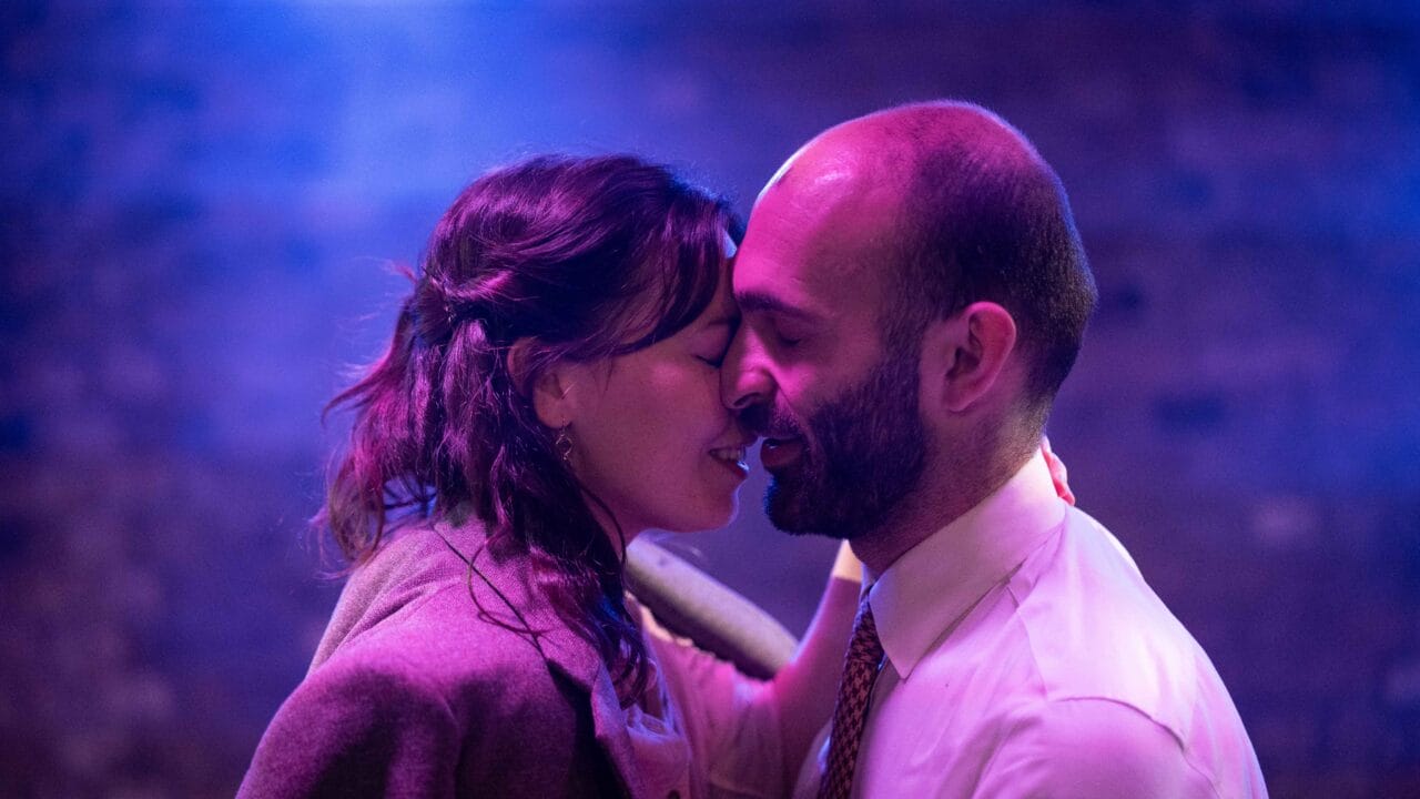 Two people stand close together, smiling with their eyes closed, as if about to kiss, in a softly lit setting with a purple and blue background.