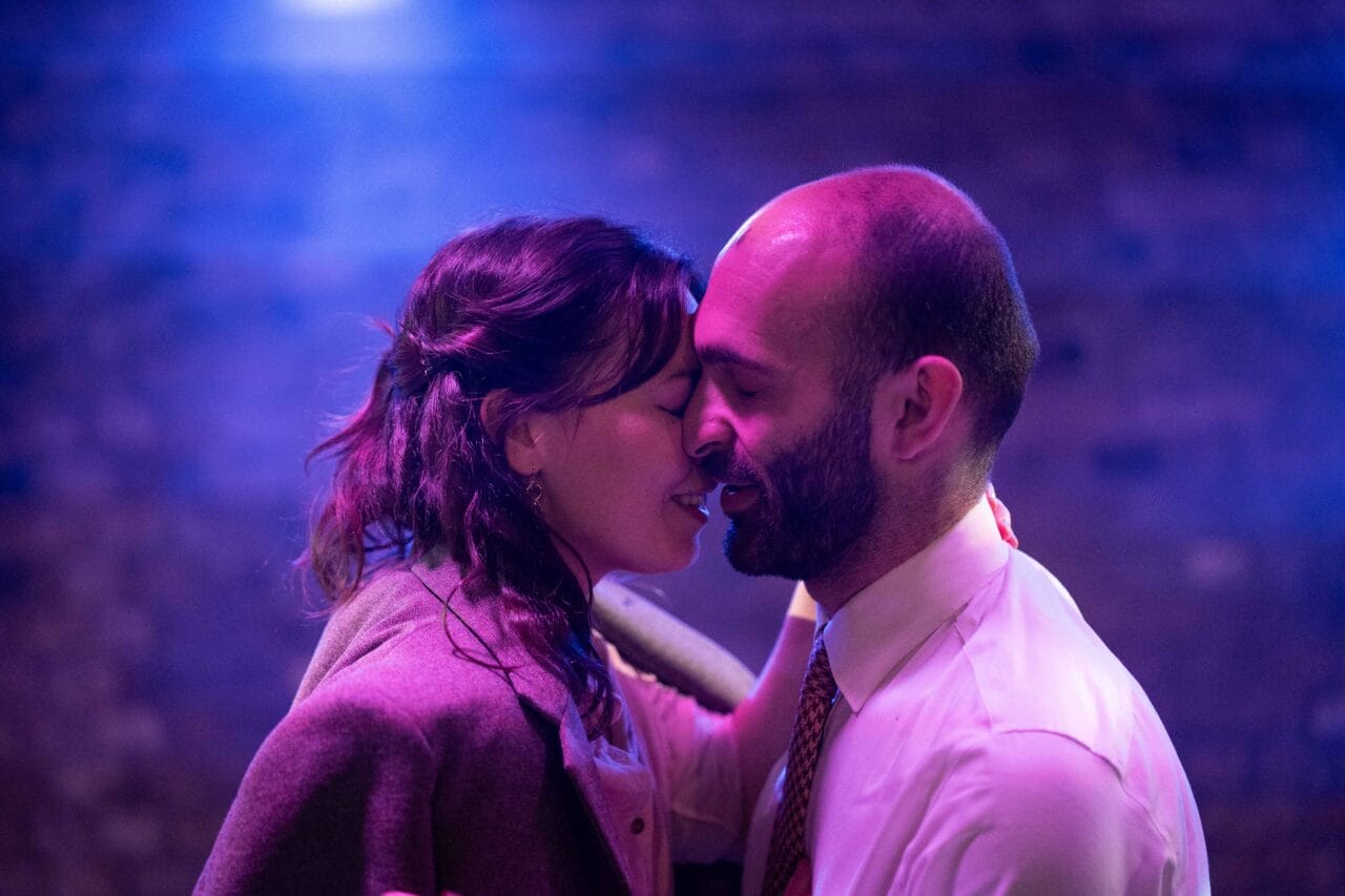 Two people stand close together, smiling with their eyes closed, as if about to kiss, in a softly lit setting with a purple and blue background.