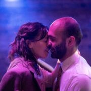 Two people stand close together, smiling with their eyes closed, as if about to kiss, in a softly lit setting with a purple and blue background.
