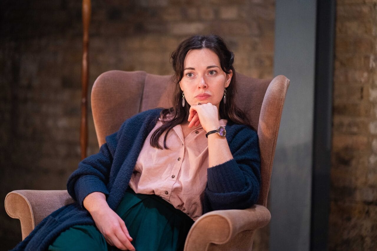 A woman sits in a brown armchair with her chin resting on her hand, wearing a pink blouse, green pants, and a dark cardigan, looking thoughtfully ahead.