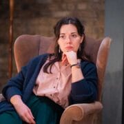 A woman sits in a brown armchair with her chin resting on her hand, wearing a pink blouse, green pants, and a dark cardigan, looking thoughtfully ahead.