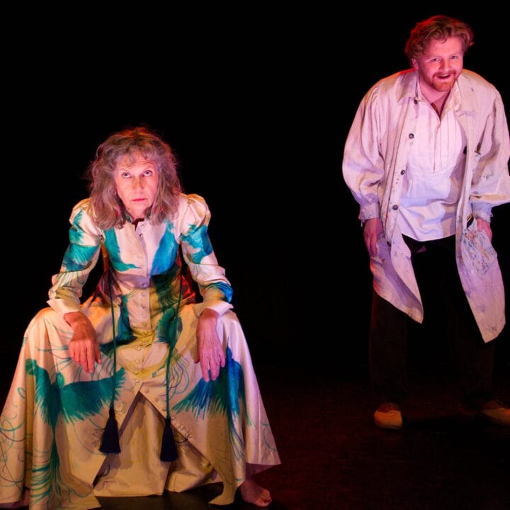 An older woman in a patterned dress sits with elbows on knees while a bearded man in a loose shirt and coat stands behind her, both on a dark stage.