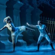 Two male ballet dancers perform on stage, one in a feathered costume posing on one leg, the other in white attire reaching out, with dramatic lighting and classical set elements.