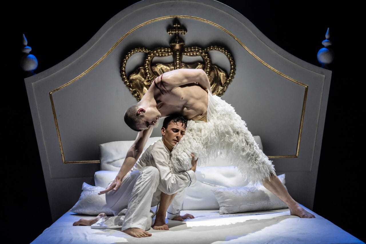 Two male dancers perform an expressive pose on a white bed with a large ornate gold crown headboard; one wears feathery white attire, the other wears plain white clothing.