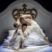 Two male dancers perform an expressive pose on a white bed with a large ornate gold crown headboard; one wears feathery white attire, the other wears plain white clothing.