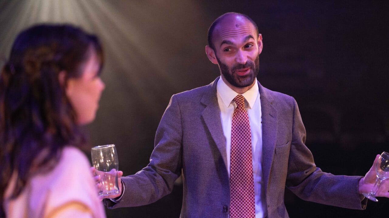 A man in a suit and tie gestures while holding a drink, speaking to a woman holding a glass in a dimly lit setting.