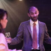 A man in a suit and tie gestures while holding a drink, speaking to a woman holding a glass in a dimly lit setting.
