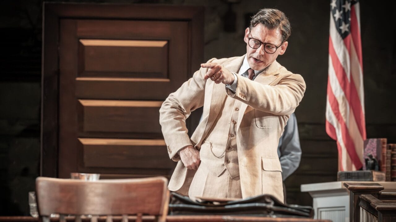 A man in a beige suit and glasses stands beside a desk, pointing forward with a serious expression—reminiscent of Atticus Finch from Harper Lee’s classic novel, To Kill a Mockingbird. An American flag is visible in the background.