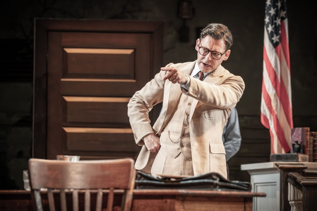 A man in a beige suit and glasses stands beside a desk, pointing forward with a serious expression—reminiscent of Atticus Finch from Harper Lee’s classic novel, To Kill a Mockingbird. An American flag is visible in the background.
