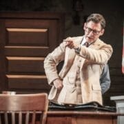 A man in a beige suit and glasses stands beside a desk, pointing forward with a serious expression—reminiscent of Atticus Finch from Harper Lee’s classic novel, To Kill a Mockingbird. An American flag is visible in the background.