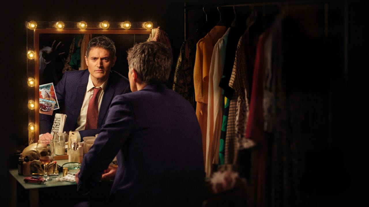 Inspector Morse, a man in a suit sits at a cluttered dressing table, looking into a mirror surrounded by lights. Clothing hangs on a rack nearby.
