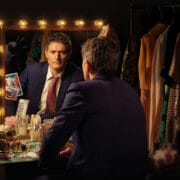 Inspector Morse, a man in a suit sits at a cluttered dressing table, looking into a mirror surrounded by lights. Clothing hangs on a rack nearby.