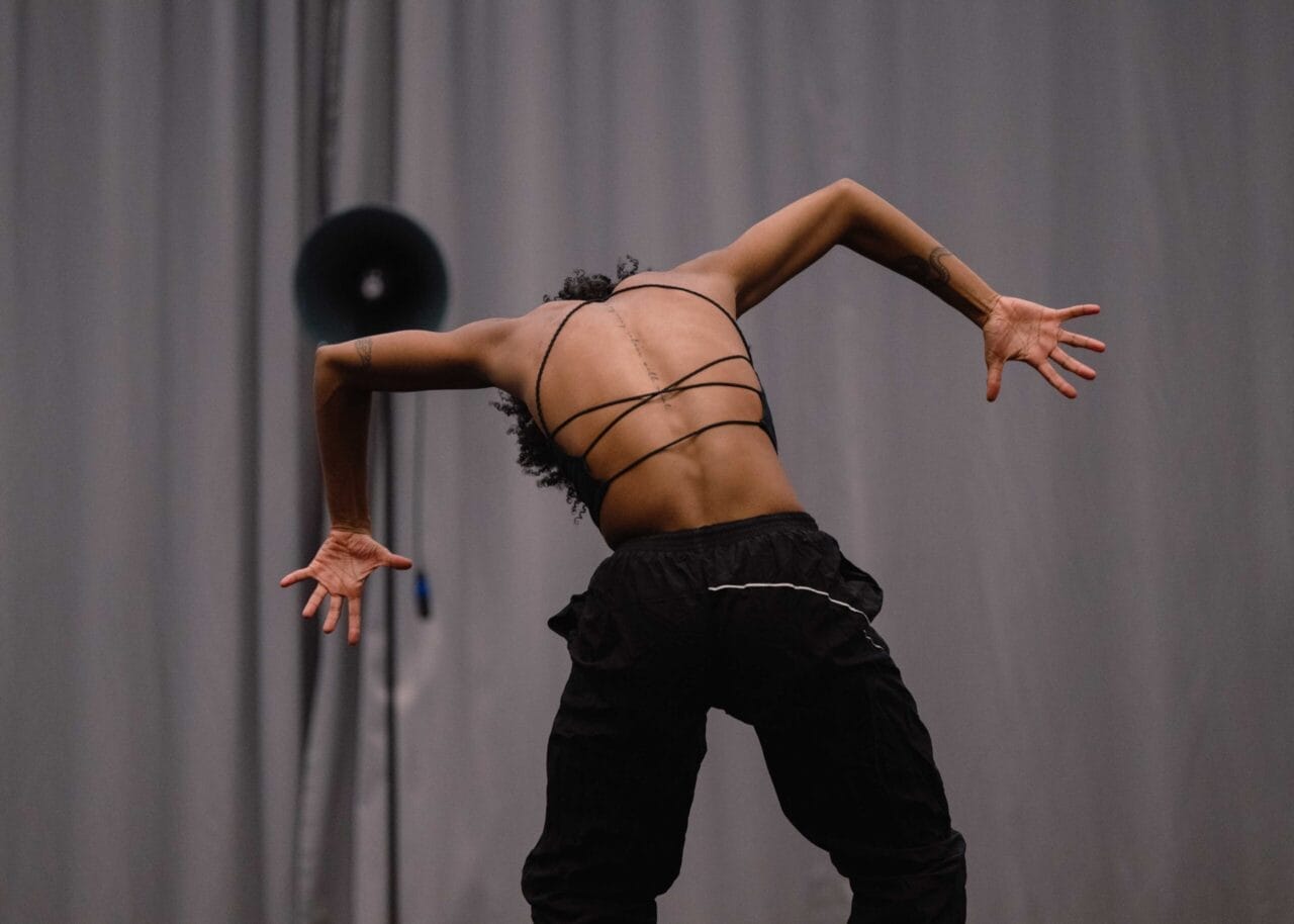 A person in a strappy top and black pants stands with their back to the camera, arms bent and fingers splayed, in front of a gray curtain and a round black speaker.