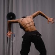 A person in a strappy top and black pants stands with their back to the camera, arms bent and fingers splayed, in front of a gray curtain and a round black speaker.