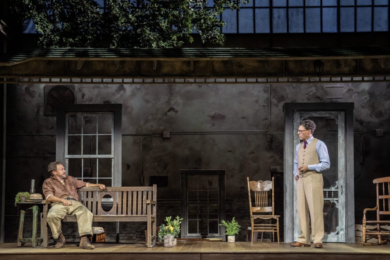 Two men perform on a stage set resembling a rustic porch, with potted plants and wooden furniture—a scene evocative of Harper Lee's classic novel, To Kill a Mockingbird, complete with a backdrop of windows and trees.