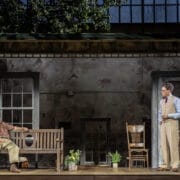 Two men perform on a stage set resembling a rustic porch, with potted plants and wooden furniture—a scene evocative of Harper Lee's classic novel, To Kill a Mockingbird, complete with a backdrop of windows and trees.