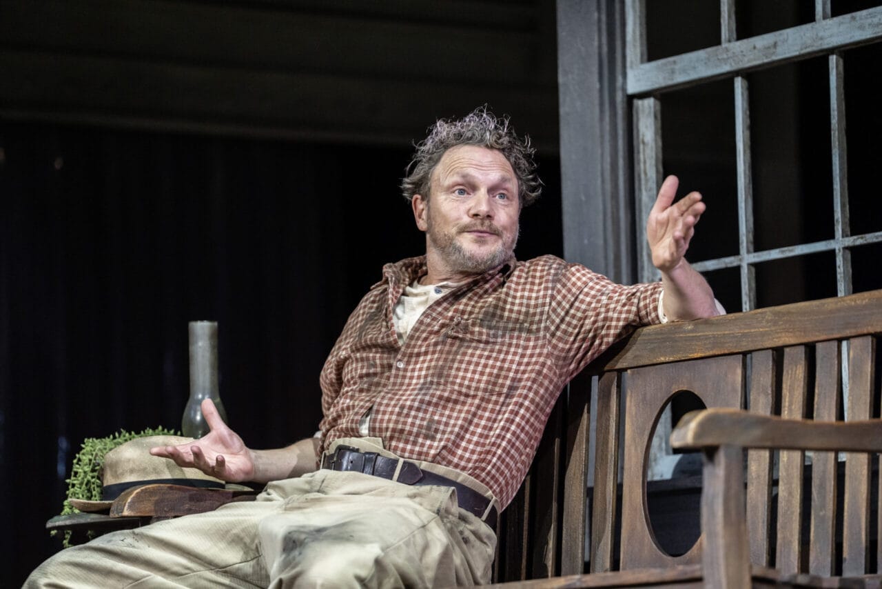 Man in a plaid shirt sits on a wooden bench indoors, gesturing with one hand while looking to the side, as if deep in thought about a classic novel like Harper Lee’s To Kill a Mockingbird. A bottle and bowl with greens are on the table beside him.
