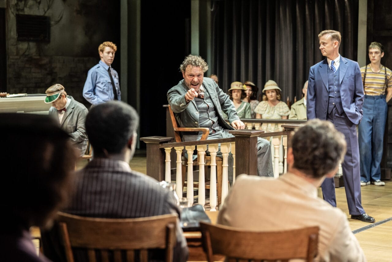 A man sits in a witness stand pointing forward during a courtroom scene, evoking the tension of Harper Lee's classic novel To Kill a Mockingbird, while others watch—including a man in a suit standing nearby.