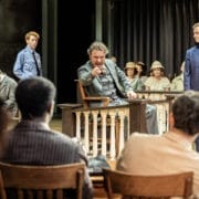 A man sits in a witness stand pointing forward during a courtroom scene, evoking the tension of Harper Lee's classic novel To Kill a Mockingbird, while others watch—including a man in a suit standing nearby.
