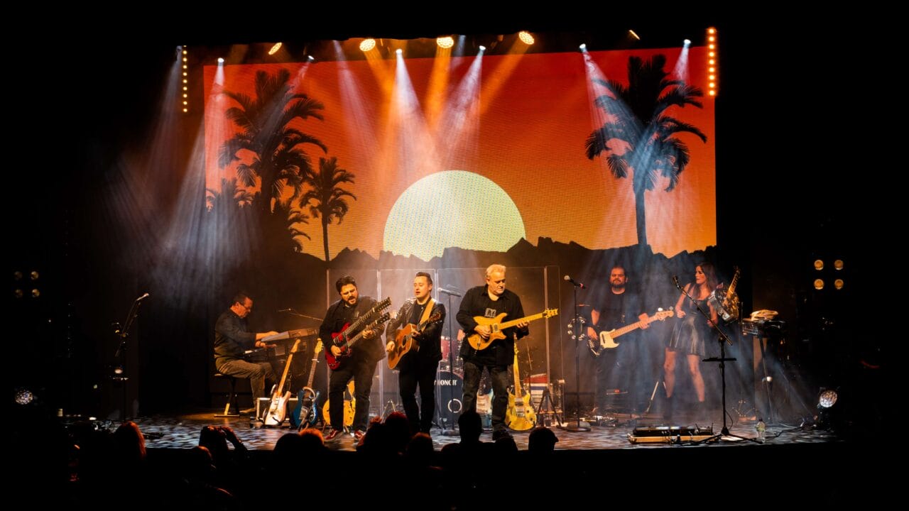 A band performs on stage with guitars and keyboards, in front of a backdrop showing a sunset with palm trees and mountains, under bright stage lights.