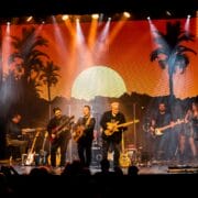 A band performs on stage with guitars and keyboards, in front of a backdrop showing a sunset with palm trees and mountains, under bright stage lights.