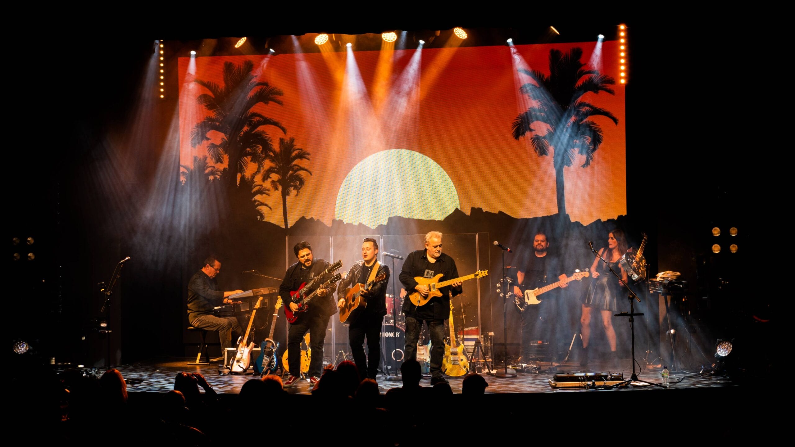 A band performs on stage with instruments under bright lights, with a sunset and palm tree backdrop projected behind them.