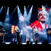 A band performs on stage under bright lights, with a large screen behind them displaying an image of a person in a royal costume and crown holding a microphone.