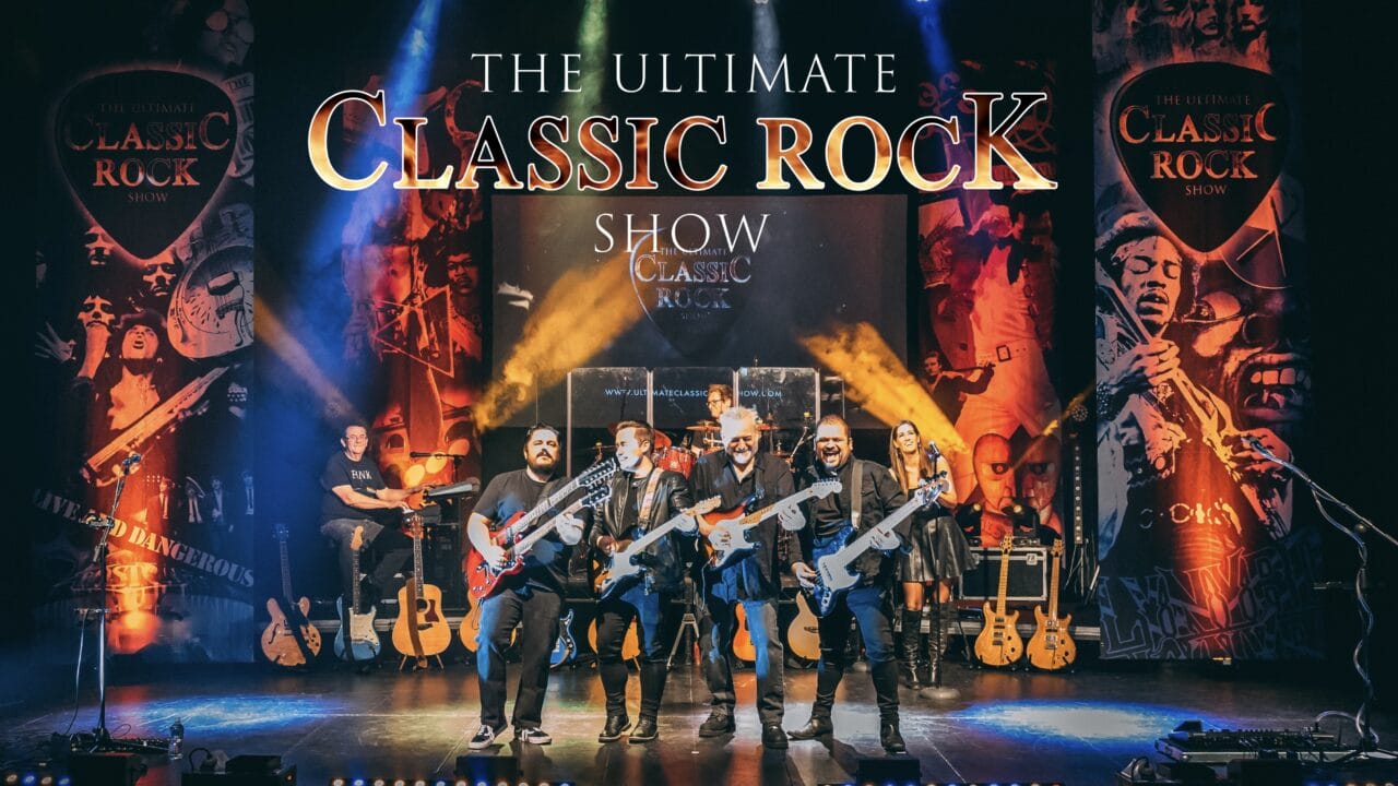 A band with guitars and a drummer performs on stage under a large sign that reads "The Ultimate Classic Rock Show," with vibrant lighting and themed banners in the background.