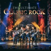 A band with guitars and a drummer performs on stage under a large sign that reads "The Ultimate Classic Rock Show," with vibrant lighting and themed banners in the background.