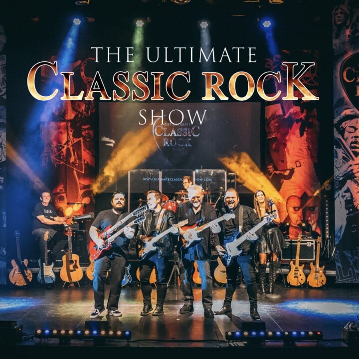 A band with guitars and a drummer performs on stage under a large sign that reads "The Ultimate Classic Rock Show," with vibrant lighting and themed banners in the background.
