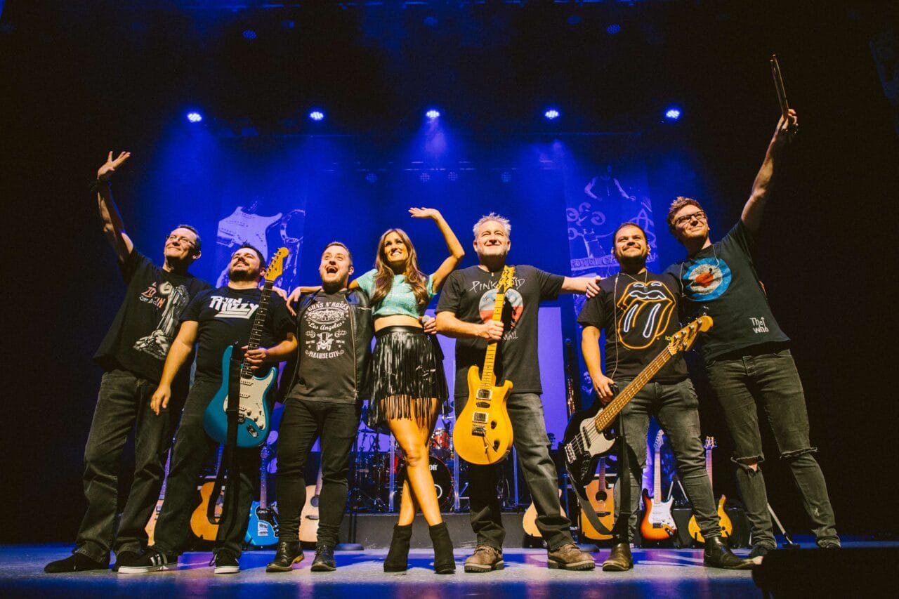 Seven band members stand on stage under blue lights, holding instruments and smiling at the audience after a performance.