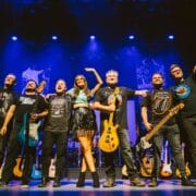 Seven band members stand on stage under blue lights, holding instruments and smiling at the audience after a performance.