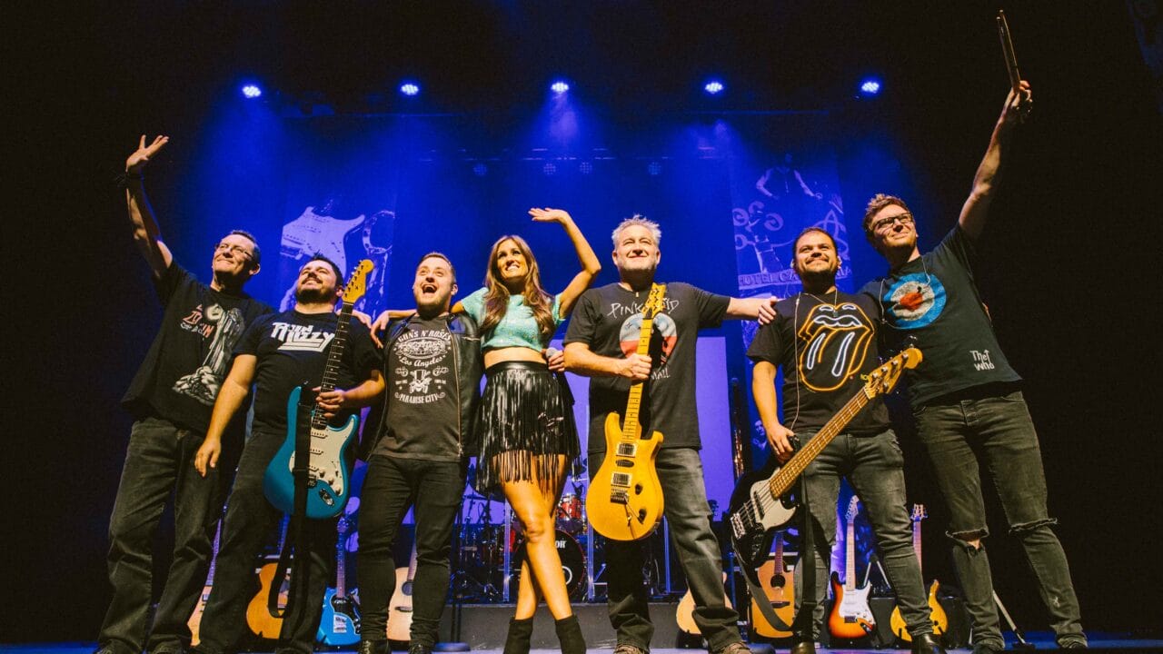 Seven musicians stand together on stage with instruments under blue lights, smiling and posing for the audience after a performance.