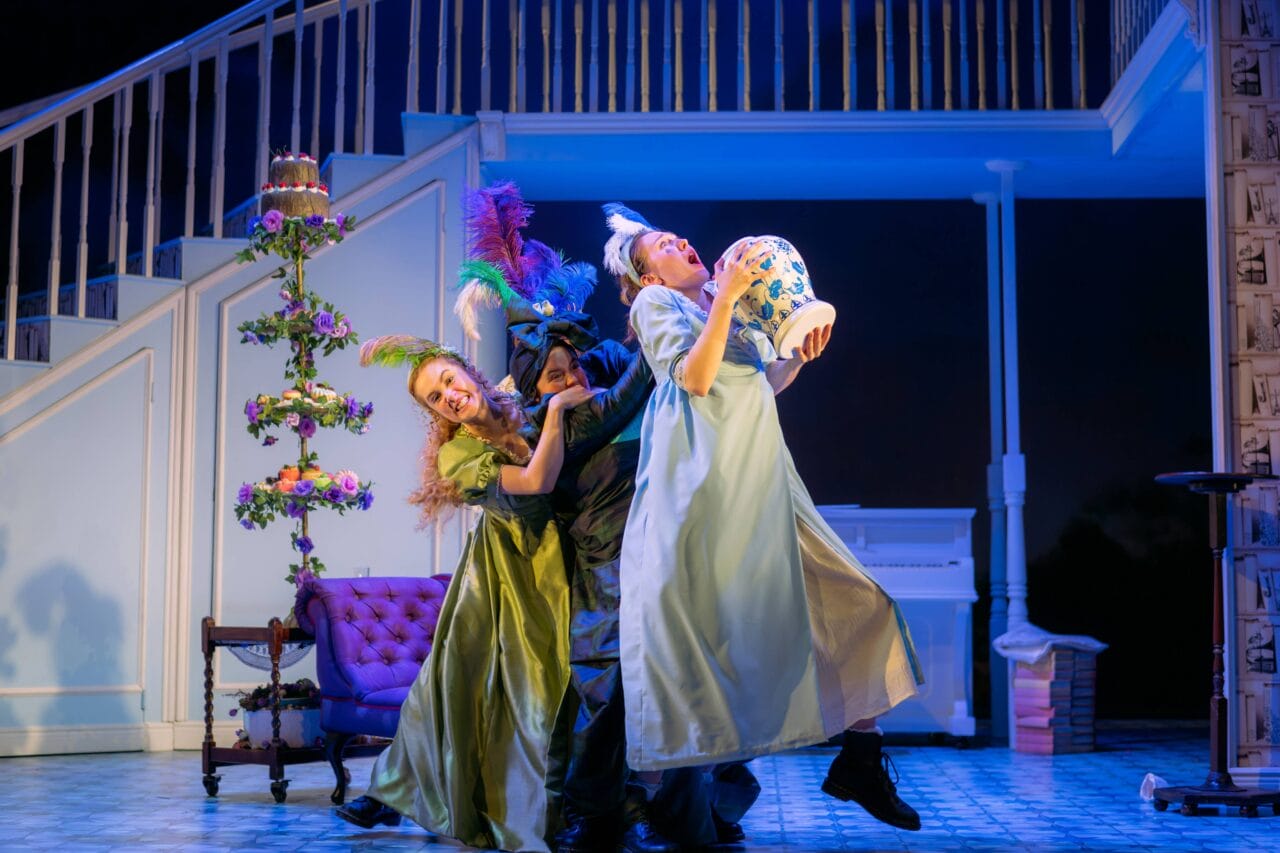 Three actors in colourful costumes perform a dramatic scene onstage; one holds a cake, and the background features a staircase, couch, and piano.