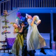 Three actors in colourful costumes perform a dramatic scene onstage; one holds a cake, and the background features a staircase, couch, and piano.