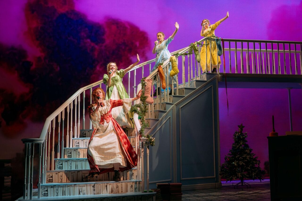 Four actors in period costumes sing and gesture on an ornate staircase set, with a dramatic purple sky backdrop and a decorated Christmas tree visible below.