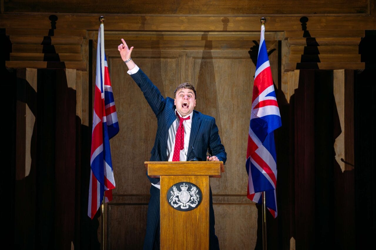 A man in a suit speaks energetically at a podium between two Union Jack flags, raising one arm upward in a wooden-paneled room.