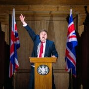 A man in a suit speaks energetically at a podium between two Union Jack flags, raising one arm upward in a wooden-paneled room.