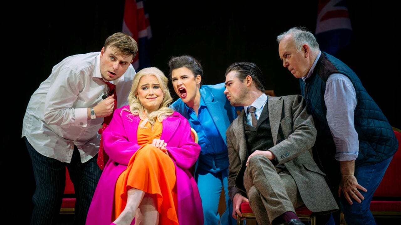 Five actors on stage, one woman in bright pink and orange sits center, while four others stand or lean around her, all with expressive faces; UK flags are visible in the background.