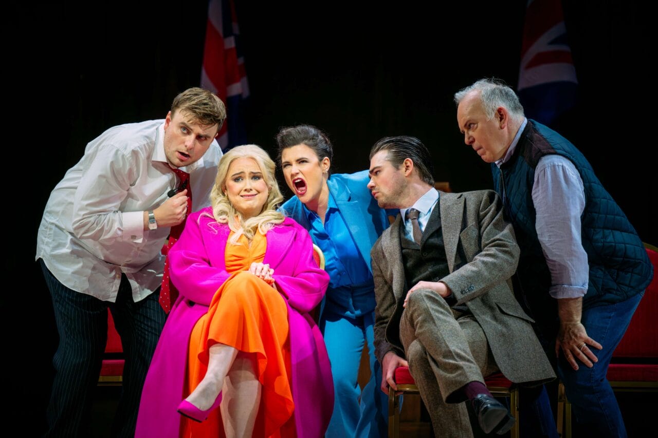 Five actors on stage, one woman in bright pink and orange sits center, while four others stand or lean around her, all with expressive faces; UK flags are visible in the background.