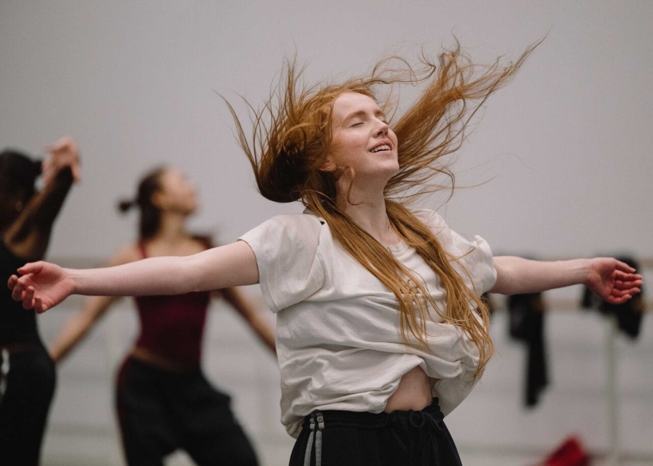 A woman with long red hair dances with eyes closed and arms outstretched in a studio, with other dancers blurred in the background.