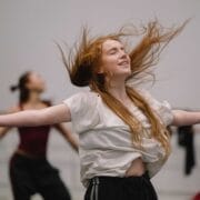 A woman with long red hair dances with eyes closed and arms outstretched in a studio, with other dancers blurred in the background.