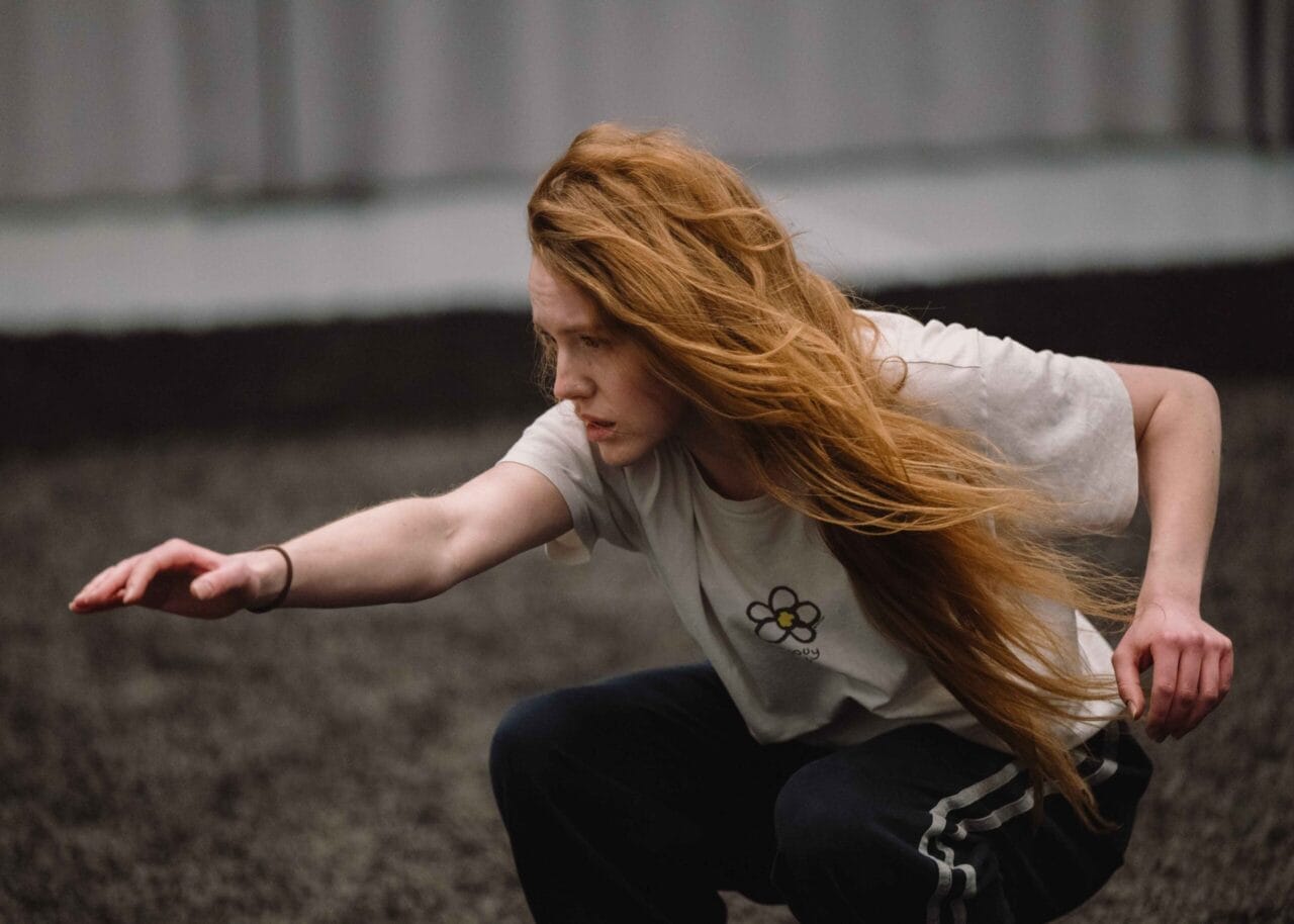 A person with long red hair crouches on the ground, extending one arm forward, wearing a white shirt and dark pants.