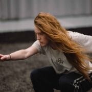 A person with long red hair crouches on the ground, extending one arm forward, wearing a white shirt and dark pants.