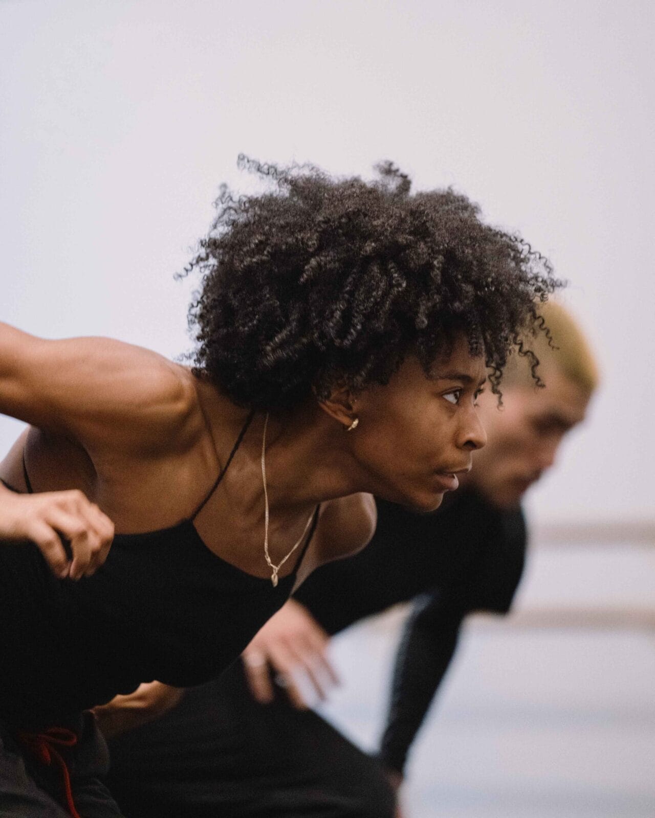 A person with curly hair in a black outfit leans forward in a dance pose, with another person blurred in the background.