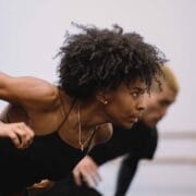 A person with curly hair in a black outfit leans forward in a dance pose, with another person blurred in the background.