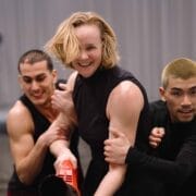 Three people, two men and one woman, stand close together indoors. The woman in the center smiles while the others hold her arms. All wear black sleeveless or short-sleeved tops.
