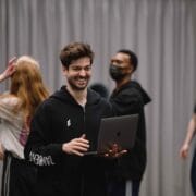 A man in a black hoodie stands smiling and holding a laptop, while several people talk and move around in a room with gray curtains.