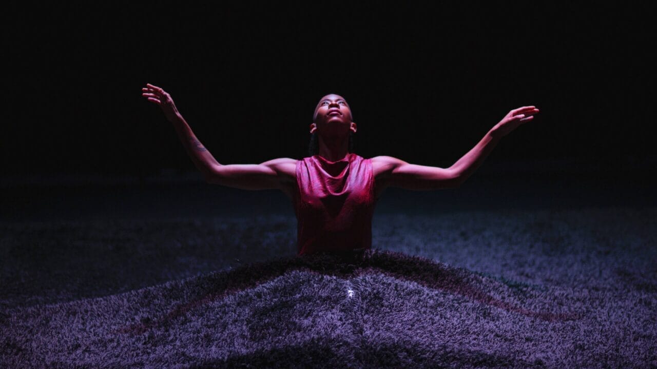 A person in a sleeveless top stands in a dark environment with arms raised, surrounded by a textured, wave-like surface illuminated by soft light.