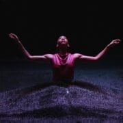 A person in a sleeveless top stands in a dark environment with arms raised, surrounded by a textured, wave-like surface illuminated by soft light.